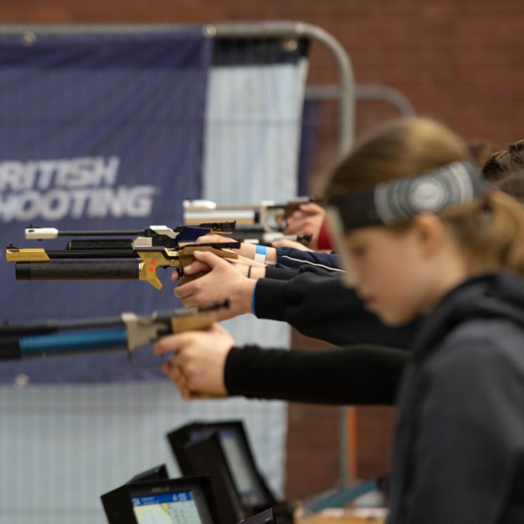 A group of young people stand at a firing point with competition air pistols. Some are aiming, some are loading. A blue banner in the background reads "British Shooting".