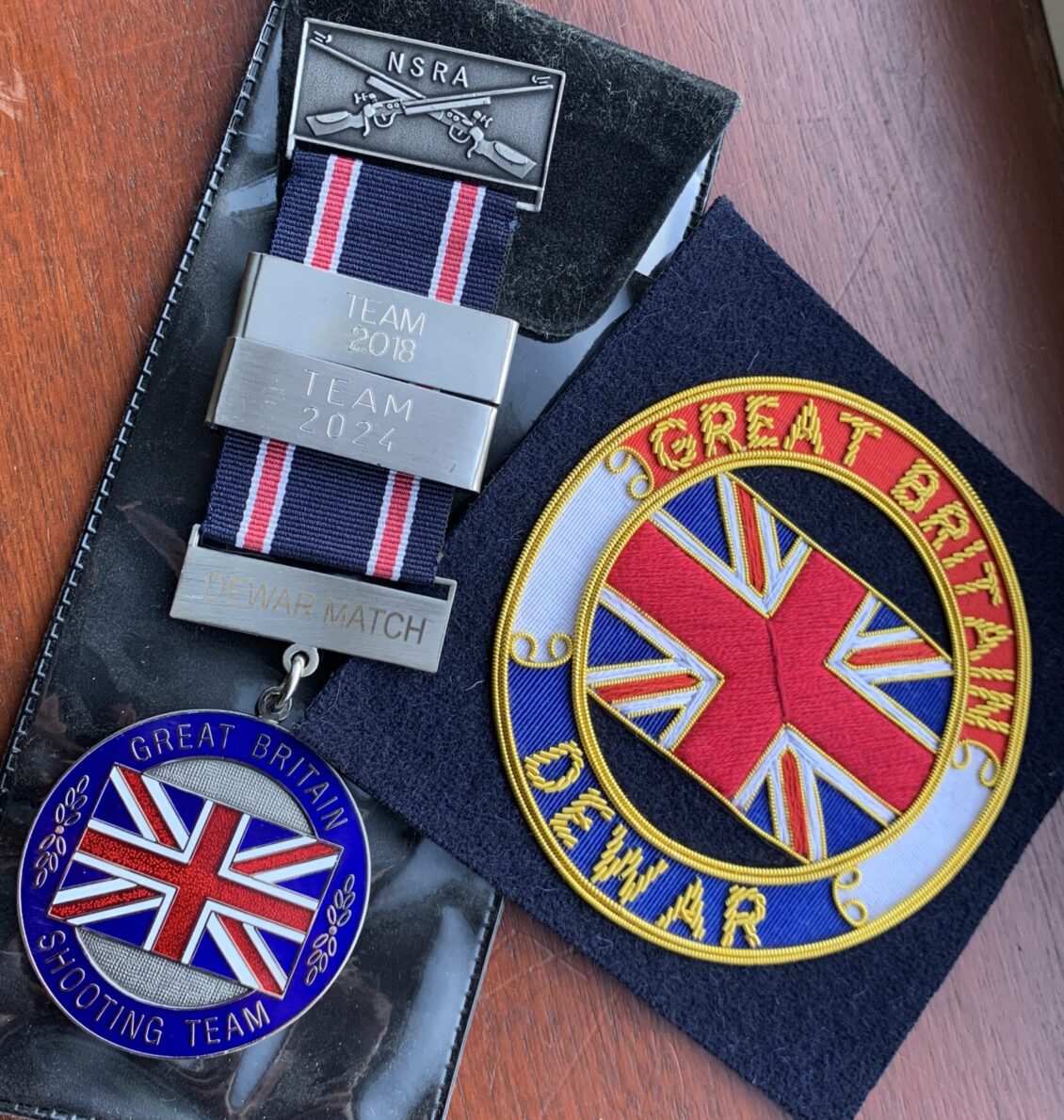 A medal for the Great Britain Shooting Team with two date bars inscribed for 2018 and 2014. Next to it is an embroidered blazer badge with the word's "Great Britain" and "Dewar".