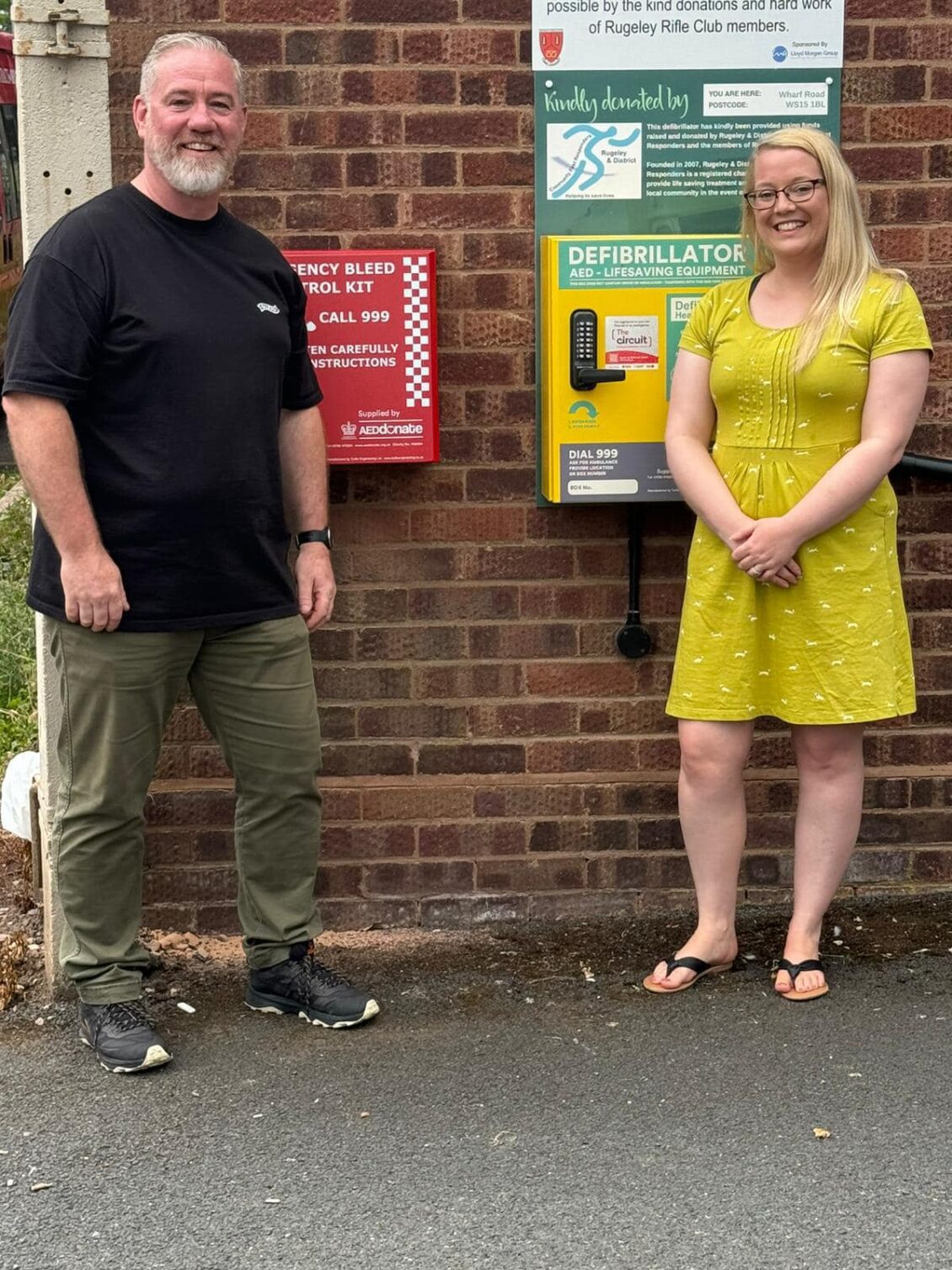 A man and a woman stand in front of a wall on which are mounted two boxes. The yellow box contains an Automatic Emergency Defibrillator. The red box contains an Emergency Bleed Control Kit