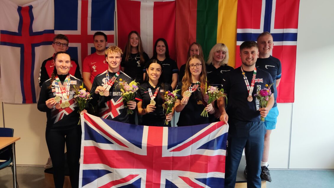 A team of smiling junior athletes stand in a group with a Union Flag in front of them at the Nordic Championships.