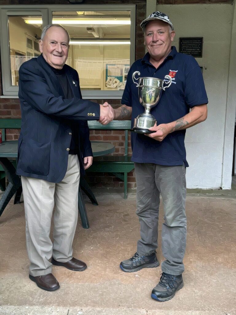 Two men stand on a veranda, the one on the right receives a trophy from the suited gentleman on the left. They are smiling and shaking hands.