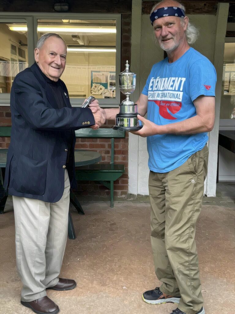 Two men stand on a veranda, the one on the right receives a trophy from the suited gentleman on the left. They are smiling and shaking hands.