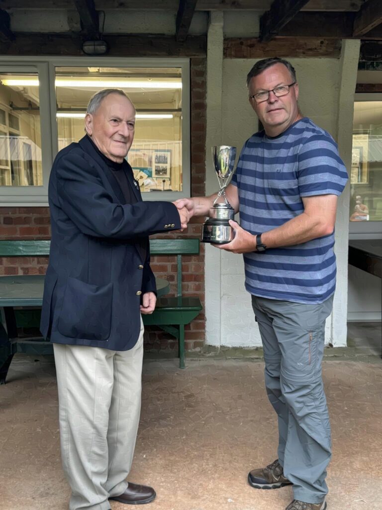 Two men stand on a veranda, the one on the right receives a trophy from the suited gentleman on the left. They are smiling and shaking hands.