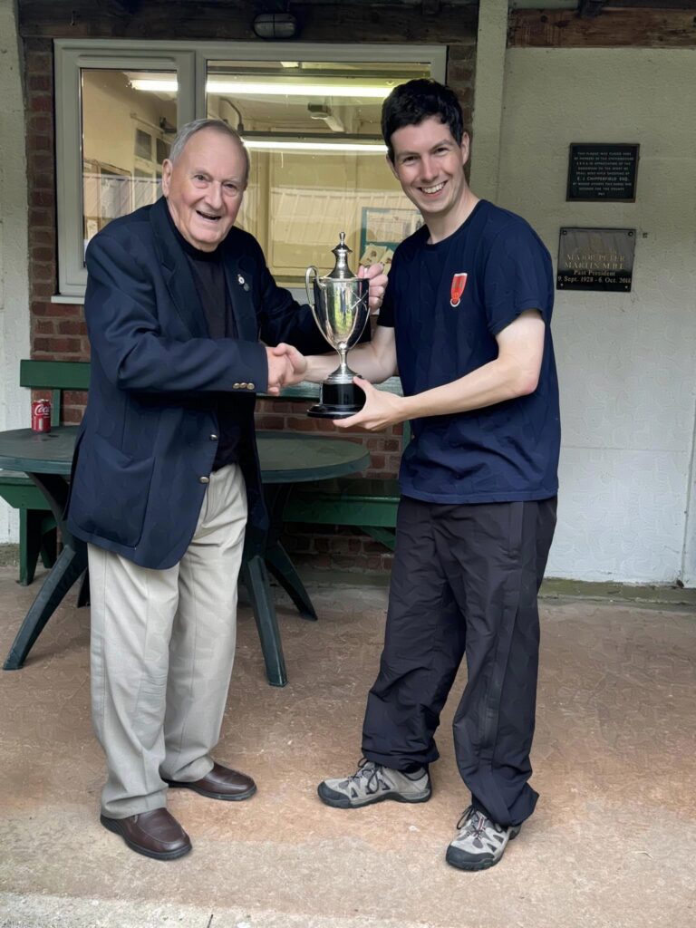 Two men stand on a veranda, the one on the right receives a trophy from the suited gentleman on the left. They are smiling and shaking hands.