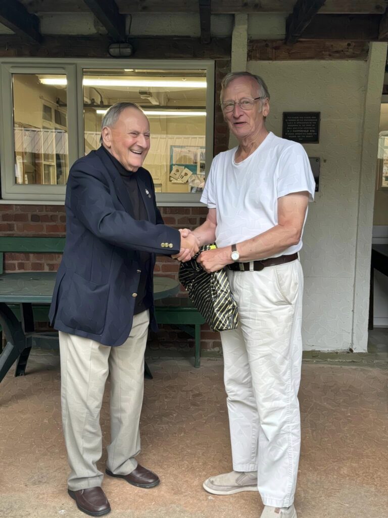 Two men stand on a veranda, the one on the right receives a trophy from the suited gentleman on the left. They are smiling and shaking hands.