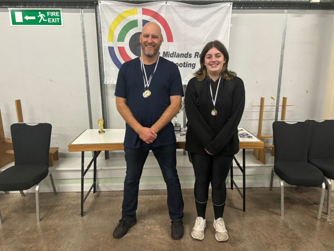 A man and a woman stand with medals indoors. A banner behind them reads "West Midlands Regional Shooting Centre".