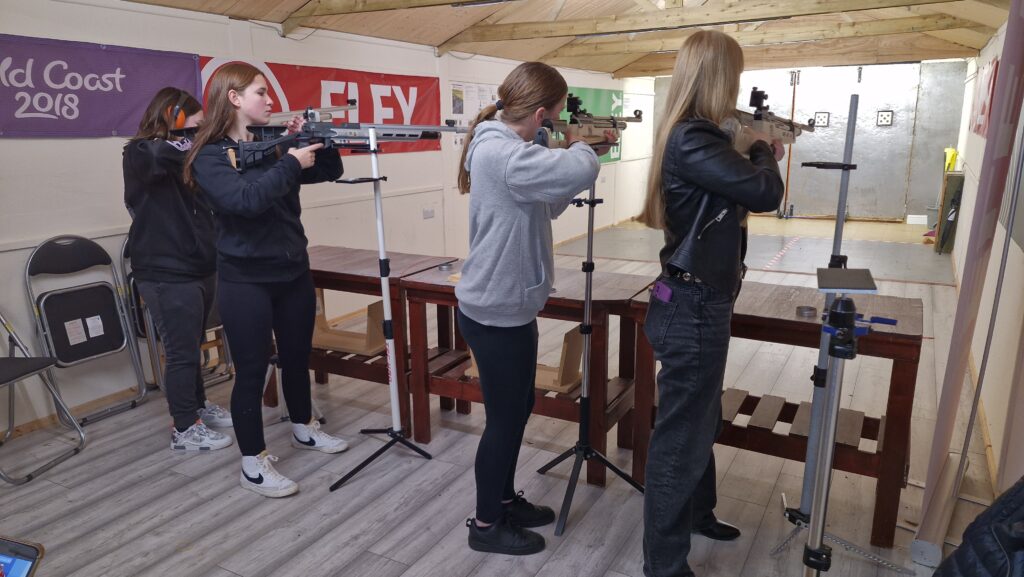 Four young women stand in an airgun range aiming target air rifles at paper targets.