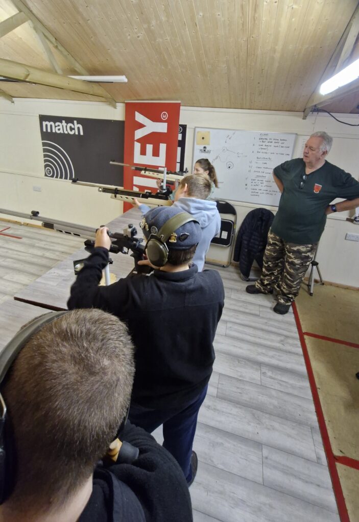 Four young people stand in an airgun range aiming target air rifles. An adult range officer supervises the firing point.