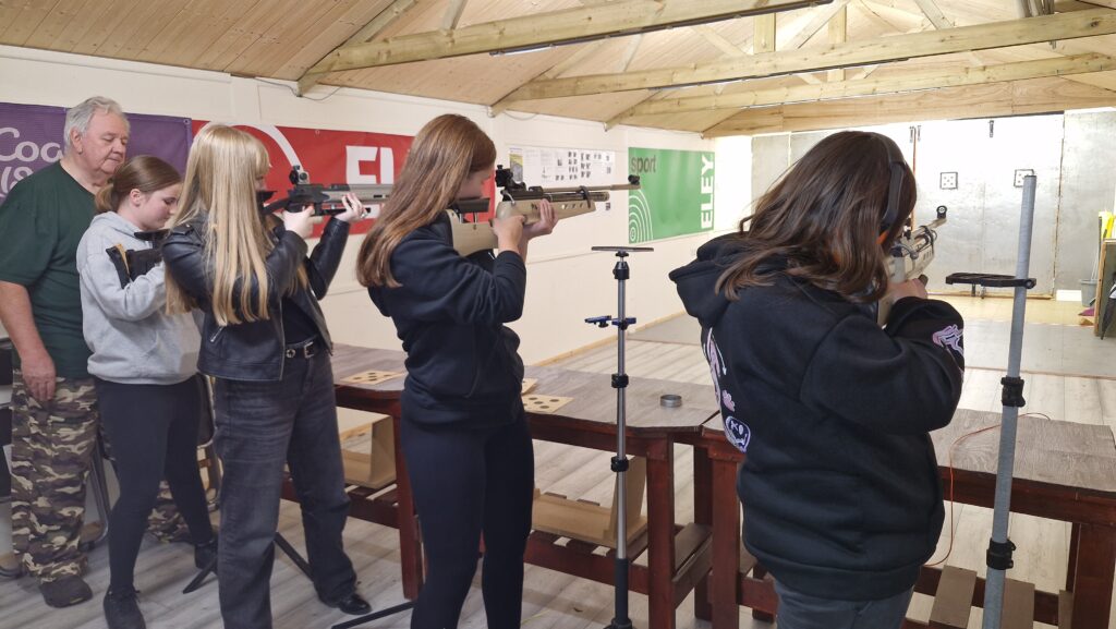 Four young women stand in an airgun range aiming target air rifles at paper targets. An adult range officer supervises the firing point.