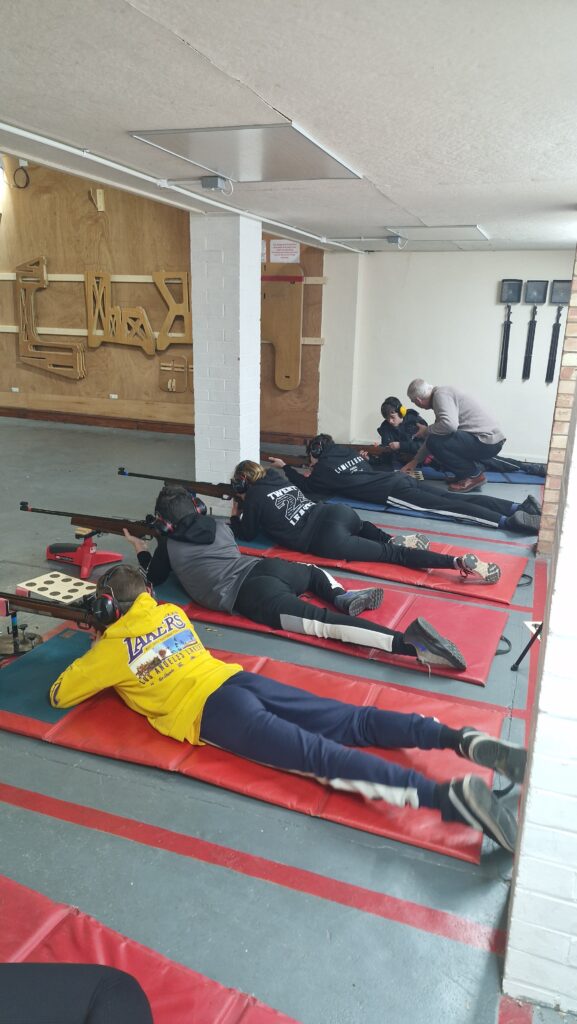 A group of young shooters aim rimfire target rifles on an indoor range. The rifles are supported at the front by stands.