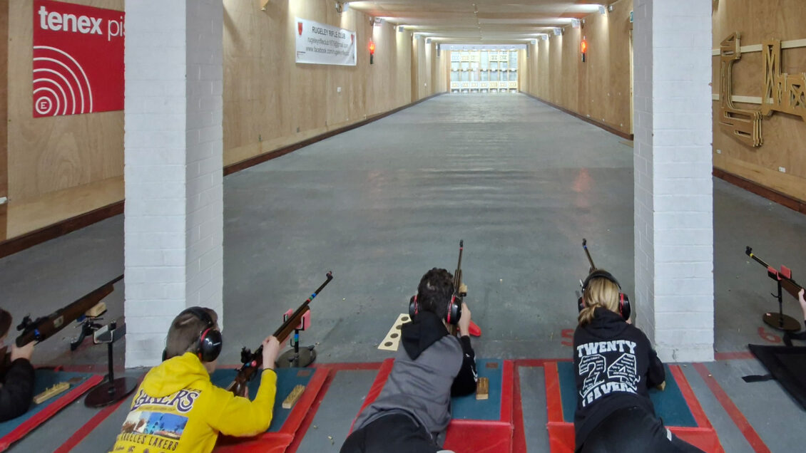 A group of young shooters aim rimfire target rifles on an indoor 25 yard range. The rifles are supported at the front by stands.