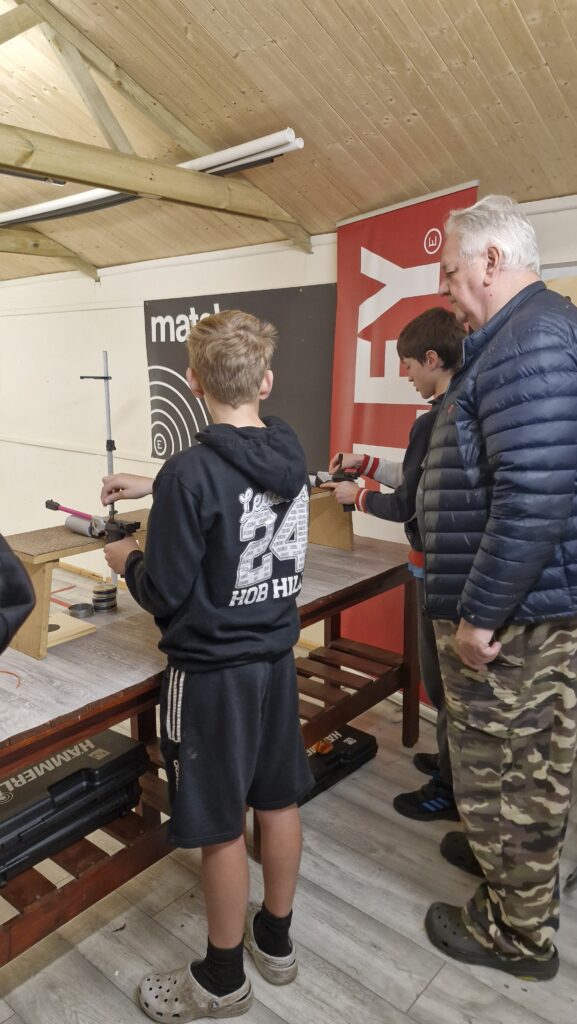 Young people on an airgun range aiming target air pistols at paper targets. An adult range officer supervises.