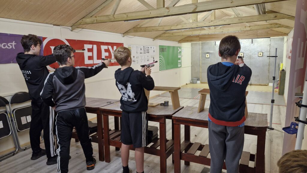 Four young men stand in an airgun range aiming target air pistols at paper targets.