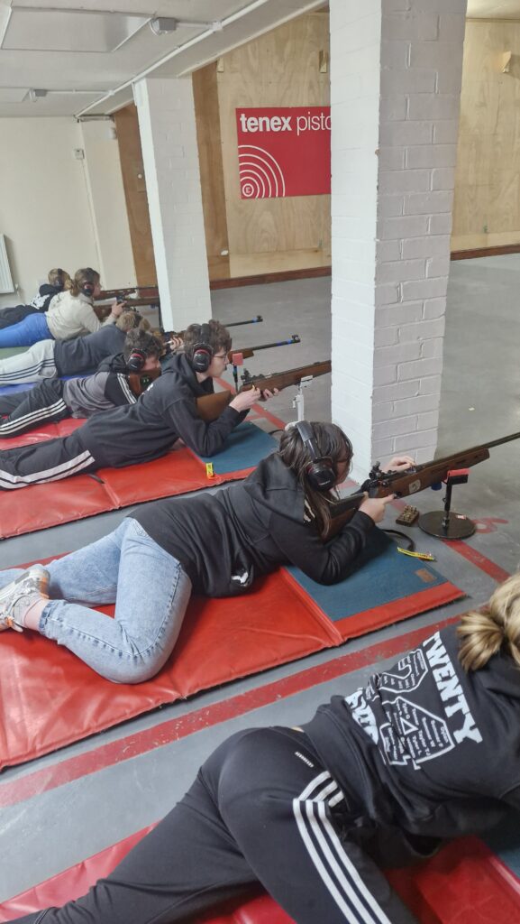 A group of young shooters aim rimfire target rifles on an indoor range. The rifles are supported at the front by stands.