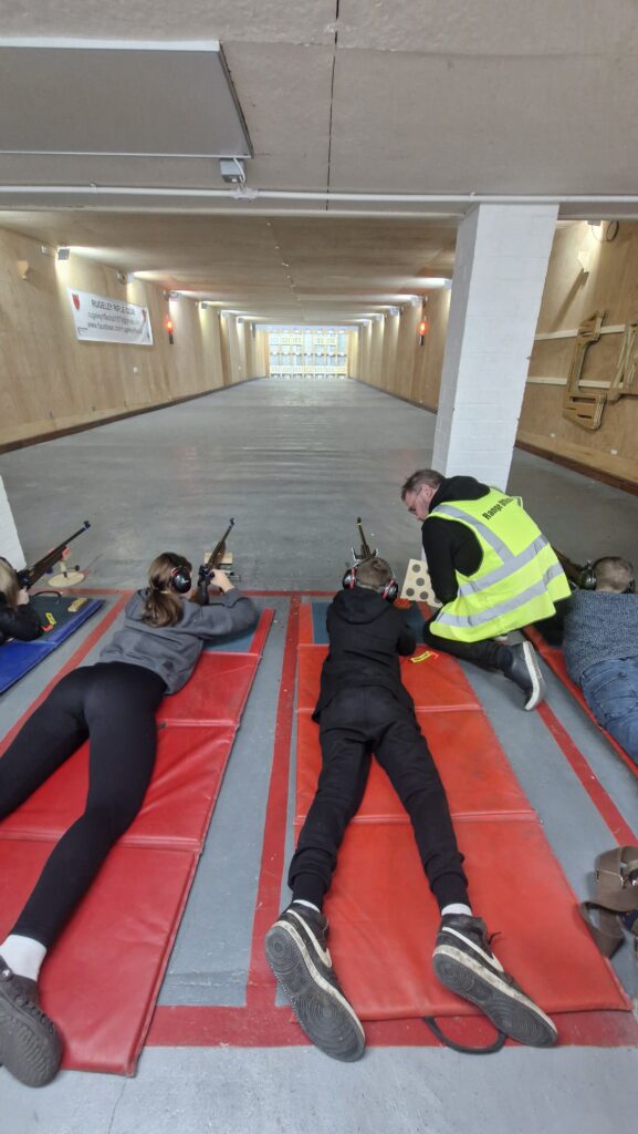 A group of young shooters aim rimfire target rifles on an indoor range. The rifles are supported at the front by stands. A coach kneels by one shooter to help them.