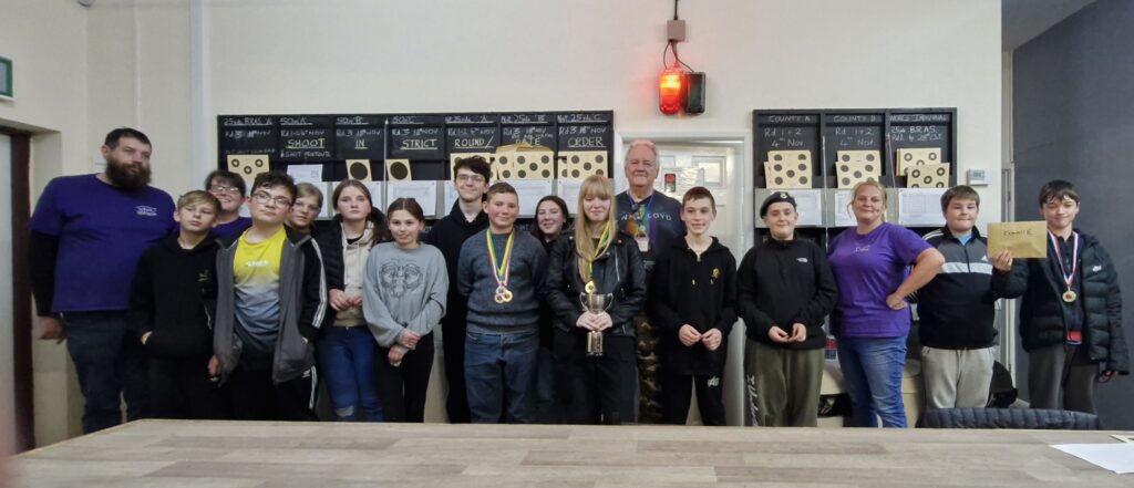 A group of young shooters stand in a clubroom with medals and prizes at the conclusion of a competition.