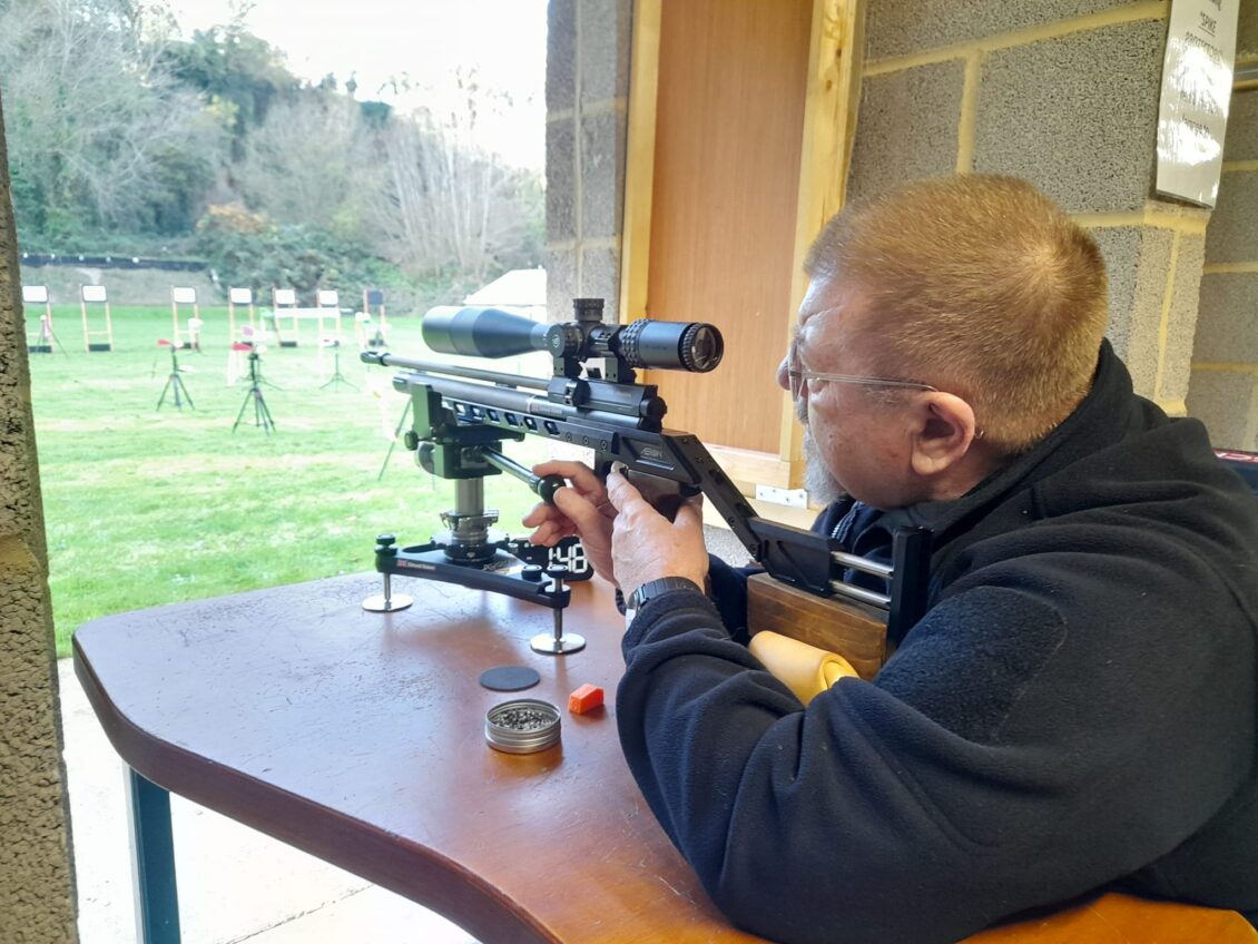 A man sits at a bench with a scoped air rifle shouldered. The air rifle sits on top of a machined rest with a fine adjusting lever to tune its point of aim.