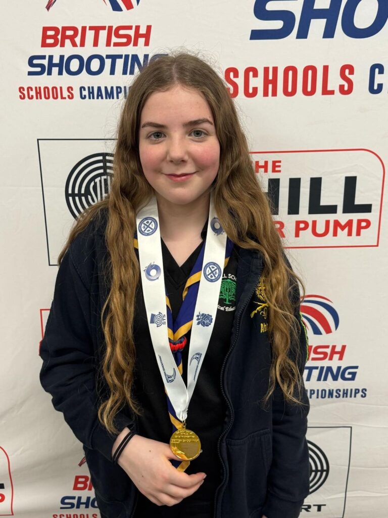 A girl stands with a gold medal in front of a backdrop reading "British Shooting Schools Championship"