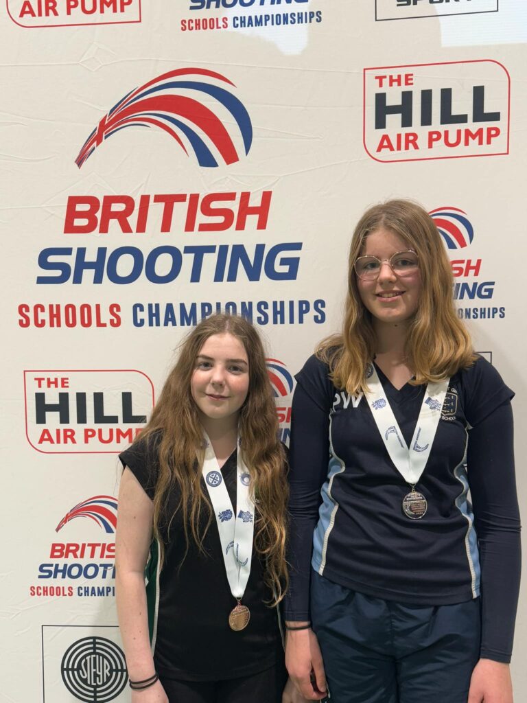 Two girls stand with medals in front of a backdrop reading "British Shooting Schools Championship"