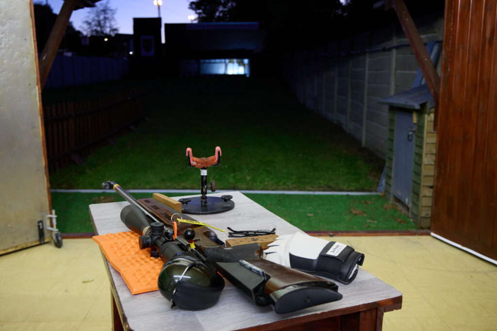 A rifle and stand lie on a bench in a building. A large door is open to the outside and targets are floodlit at the other end of the range.