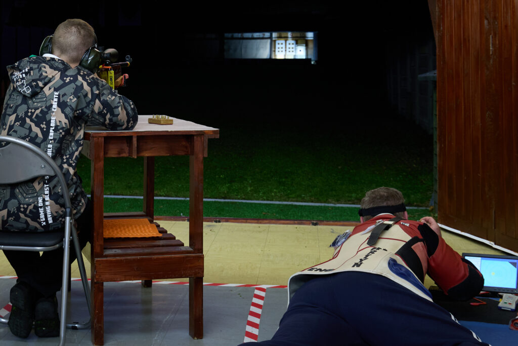 On a 50Metre outdoor range, a man aims a rifle lying down in the prone position, whilst a junior shooter aims a benchrested rifle. It is dark outside and the targets are illuminated by floodlight.