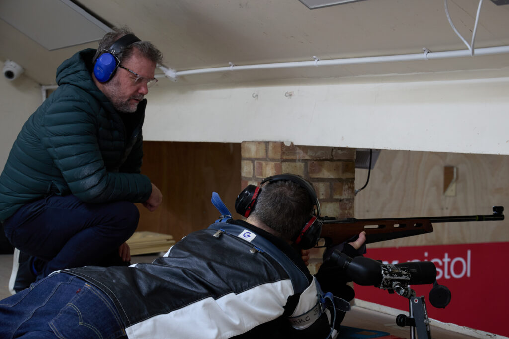 A man kneels next to a shooter holding a rifle in the prone position offering coaching and instruction.