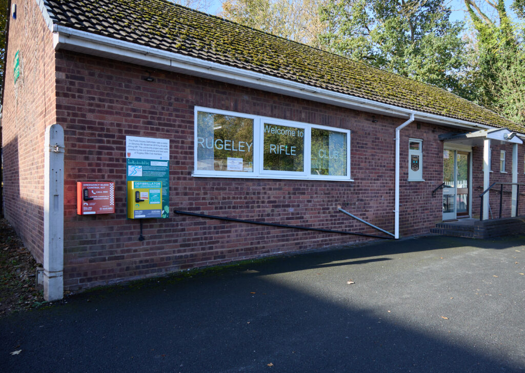 A brick building is shown. Lettering in the windows reads "Rugeley Rifle Club".

Red and yellow boxes contain an emergency bleed control kit and a public defibrillator for community use.