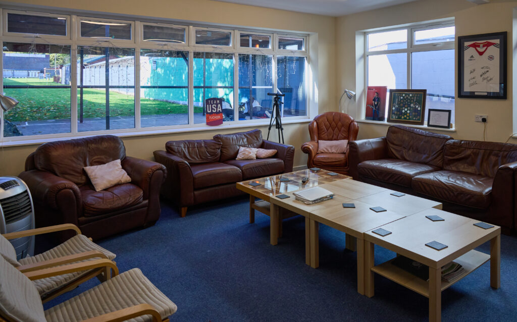 A comfortable room with big brown squishy sofas and coffee tables. A picture window fills one wall showing the view onto a firing point and outdoor rifle range. The sun is shining and two targets are visible in the distance. The room has signed t-shirts and displays of medals on the walls.