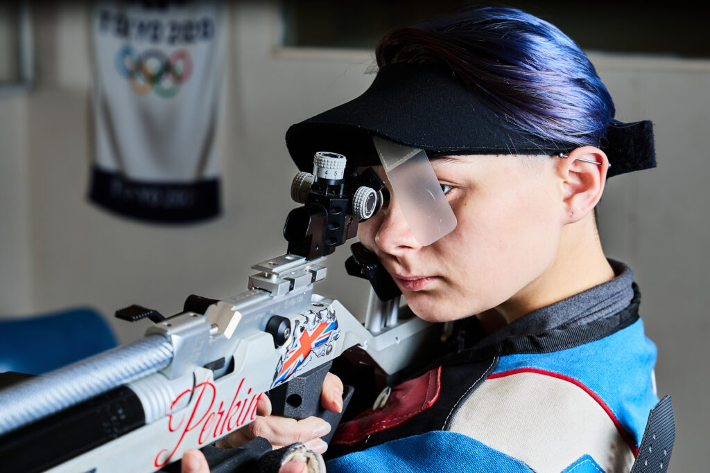 A young woman with purple hair aims a match air rifle on a shooting range. She wears a black visor to block light entering the rear of her sights, and a plastic blinder in front of her left eye to help her focus on the sight image.