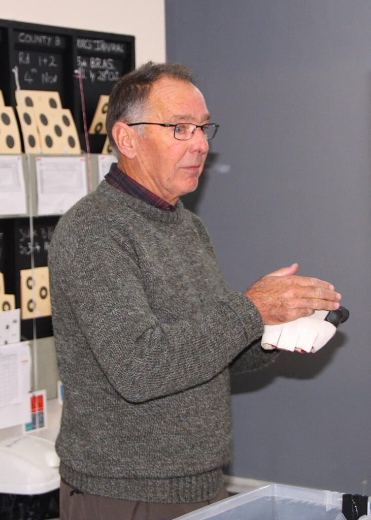 A man wearing a grey jumper stands by a table. He is holding a spacialist shooting glove as he addresses a room of shooters attending his workshop.