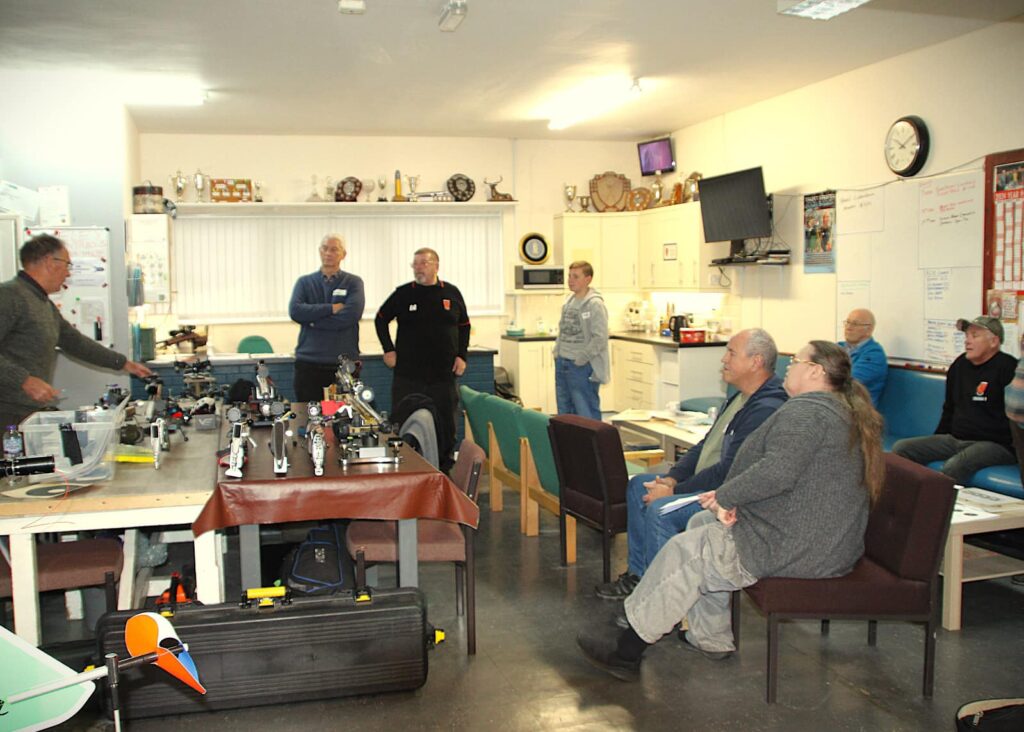 A man wearing a grey jumper stands by a table of benchrest rifles addressing a diverse group of target shooters