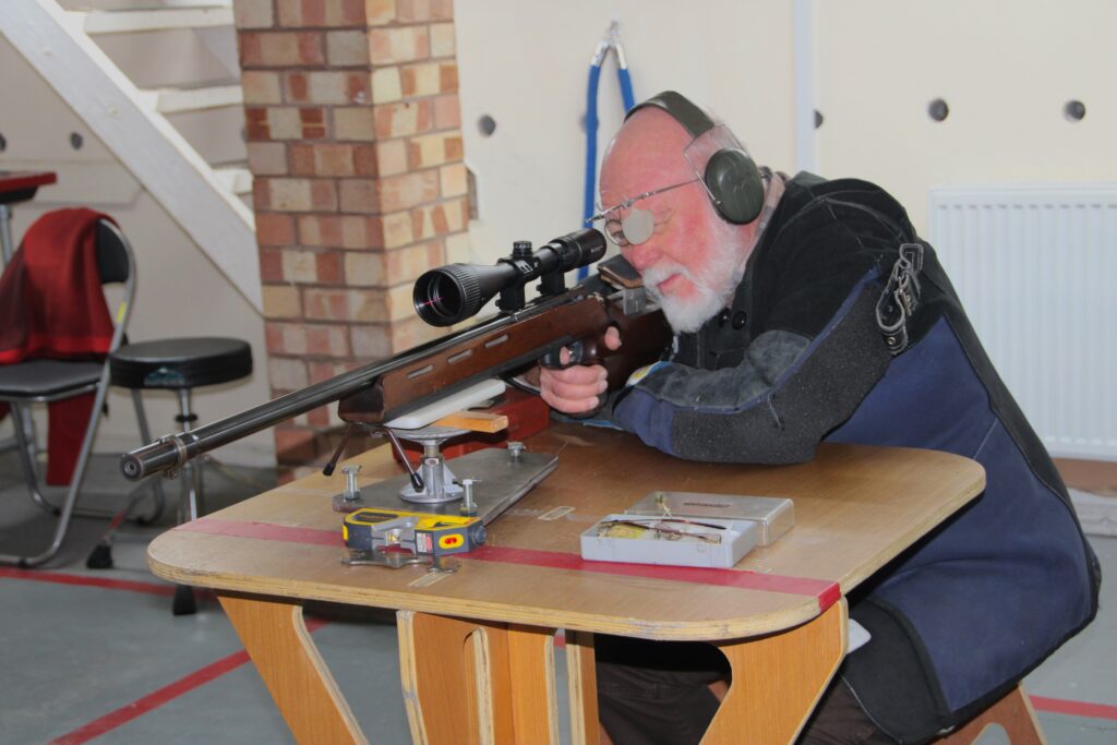 A man aims a benchrested rifle on an indoor shooting range. He wears and blue and black shooting jackets, and green ear defenders. A blinder patch covers his left eye to help him focus on the image in the scoped sight.