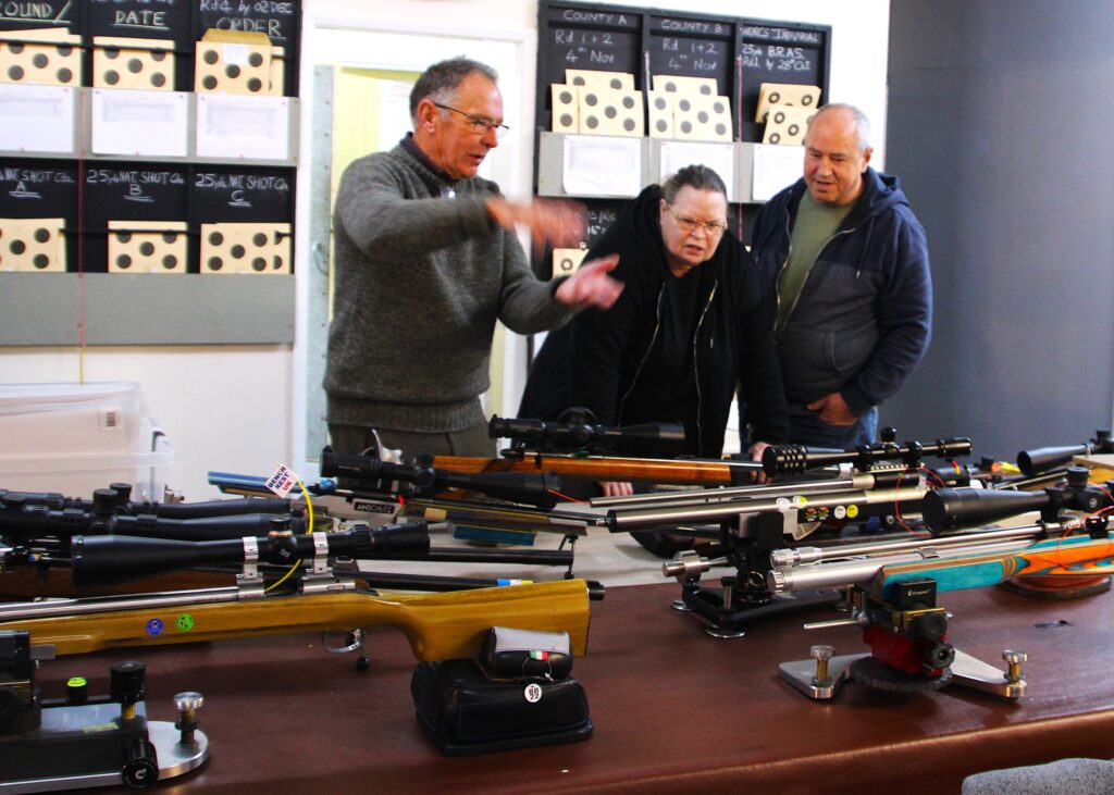 A man stands by a table of benchrest rifles discussing technique and rifles with two benchrest shooters