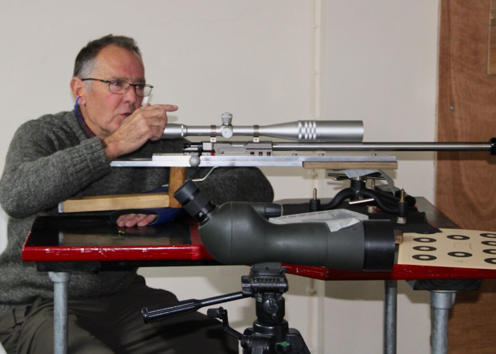 A man is sat at a table with a benchrest rifle. He is speaking and delivering a lesson on benchrest shooting.