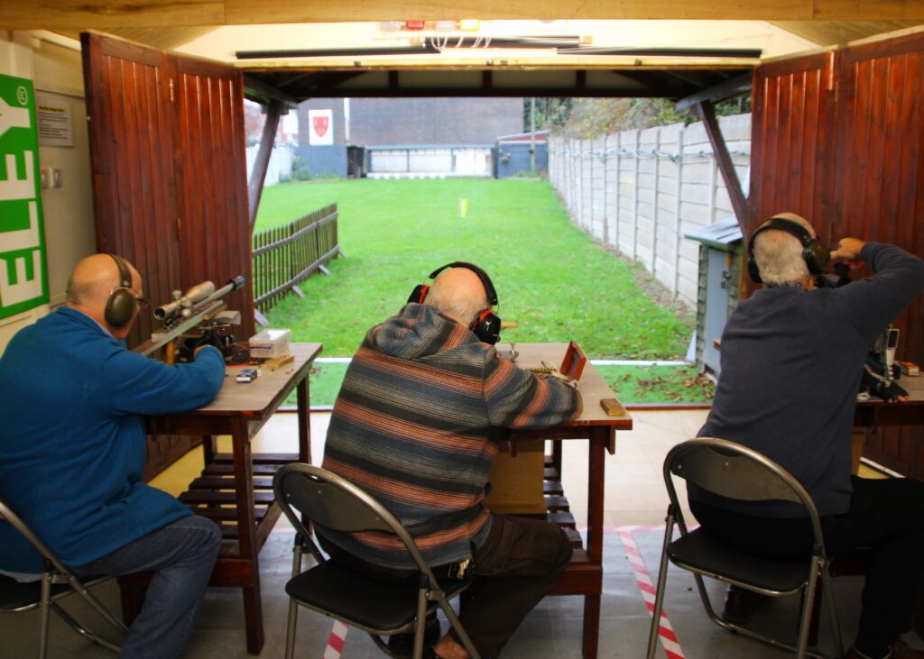 Three shooters sit an benchrest tables on a covered firing point looking out over an outdoor range.