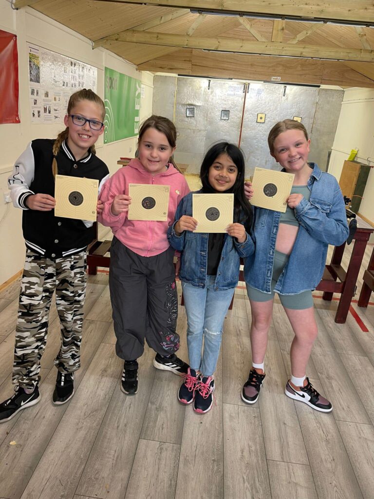 A group of four youth volunteers pose with air pistol targets on an indoor shooting range