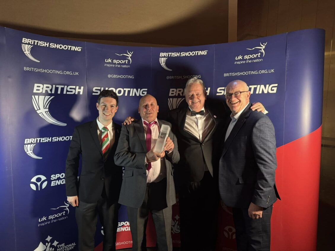 Four smiling people stand in front of a British Shooting banner. The one on the middle is holding a glass trophy.