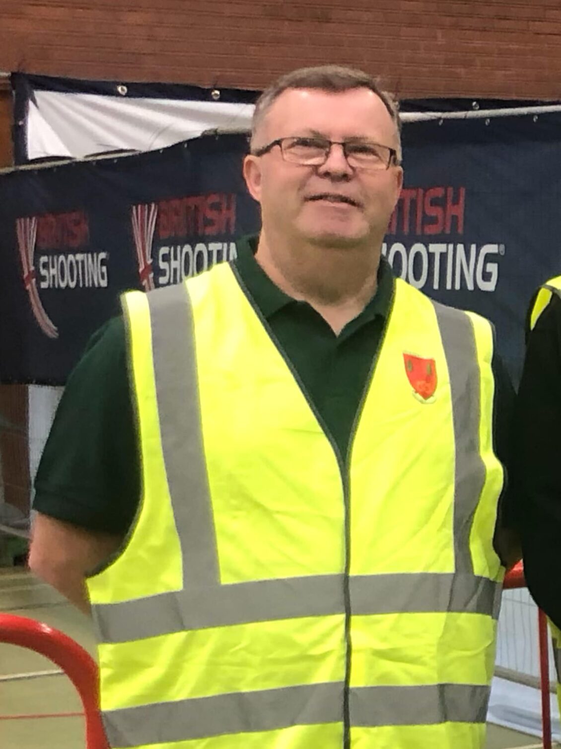 A man wearing a high-visibility jacket stands smiling in a large hall. A banner behind him reads "British Shooting"